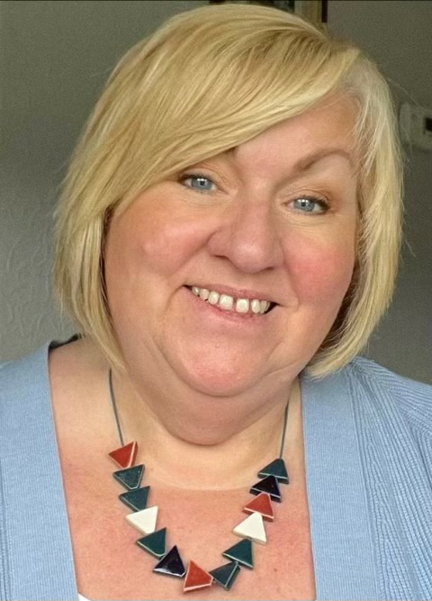 Lesley has blonde hair and is wearing a pale blue top and  has a red, white and black necklace on. Lesley is smiling.